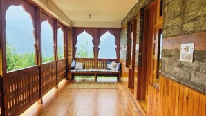 A wooden corridor with a bench, stone walls, and large windows with intricate designs. - Huts And Hunters Cottage Manali (Kullu)