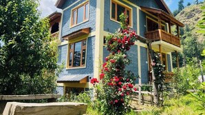 A two-story house with a balcony, surrounded by lush greenery and a garden with red flowers. - Huts And Hunters Cottage Manali (Kullu)