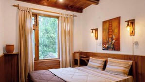 A bedroom with a large bed, wooden headboard, a window with curtains, a painting on the wall, and a small table with a chair. - Huts And Hunters Cottage Manali (Kullu)