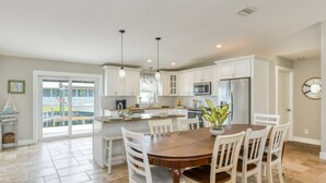 A modern kitchen with white cabinets, a central island, and a dining area with a wooden table and white chairs. - Heated Pool, Walk to Beach, 40’ Boat Dock, Kayak, Paddle Board, Fishing, $300 in Free Tickets! (St. Augustine)