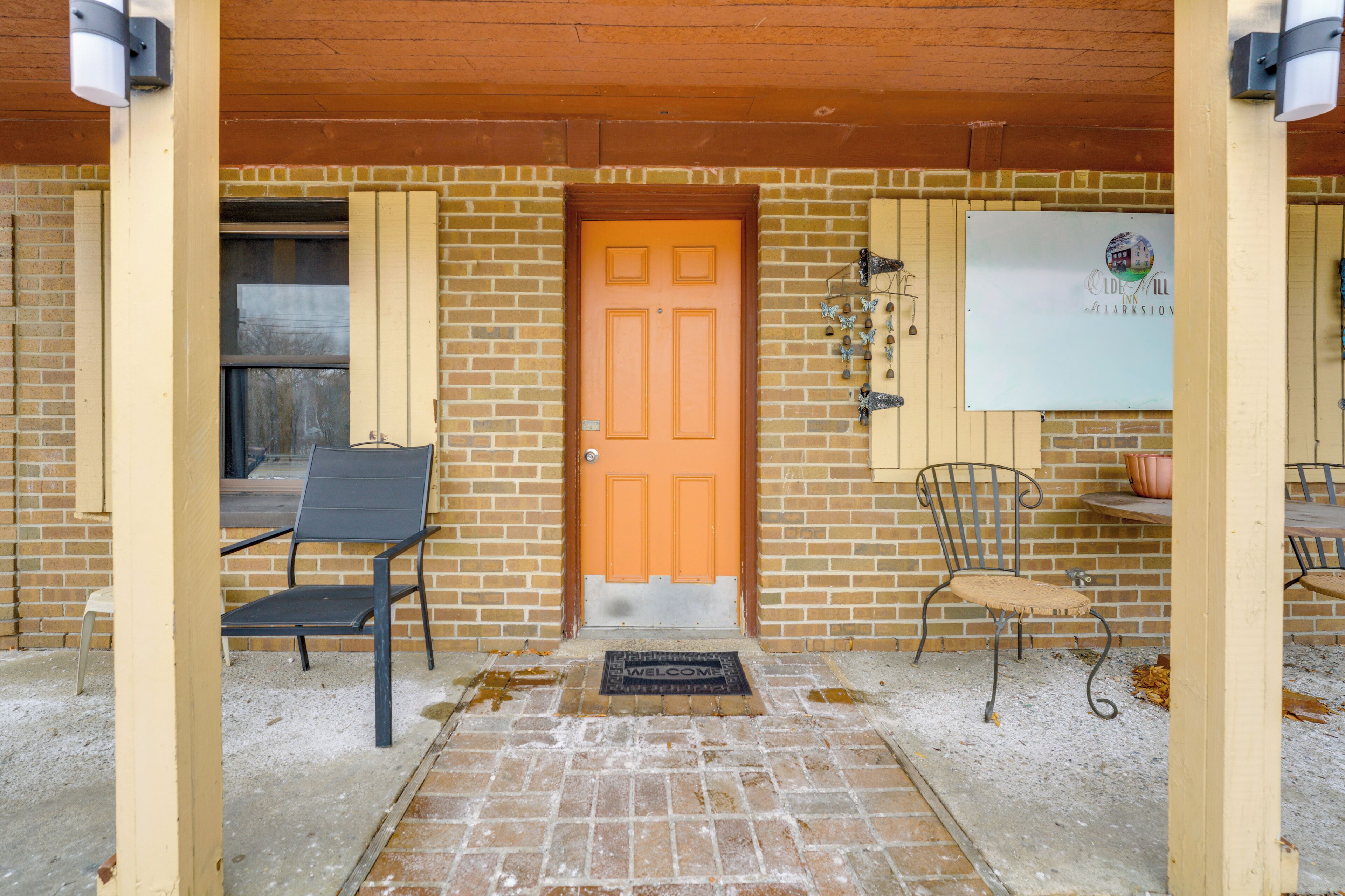 A brick building with a wooden door, two metal chairs, a welcome mat, and a sign on the wall.