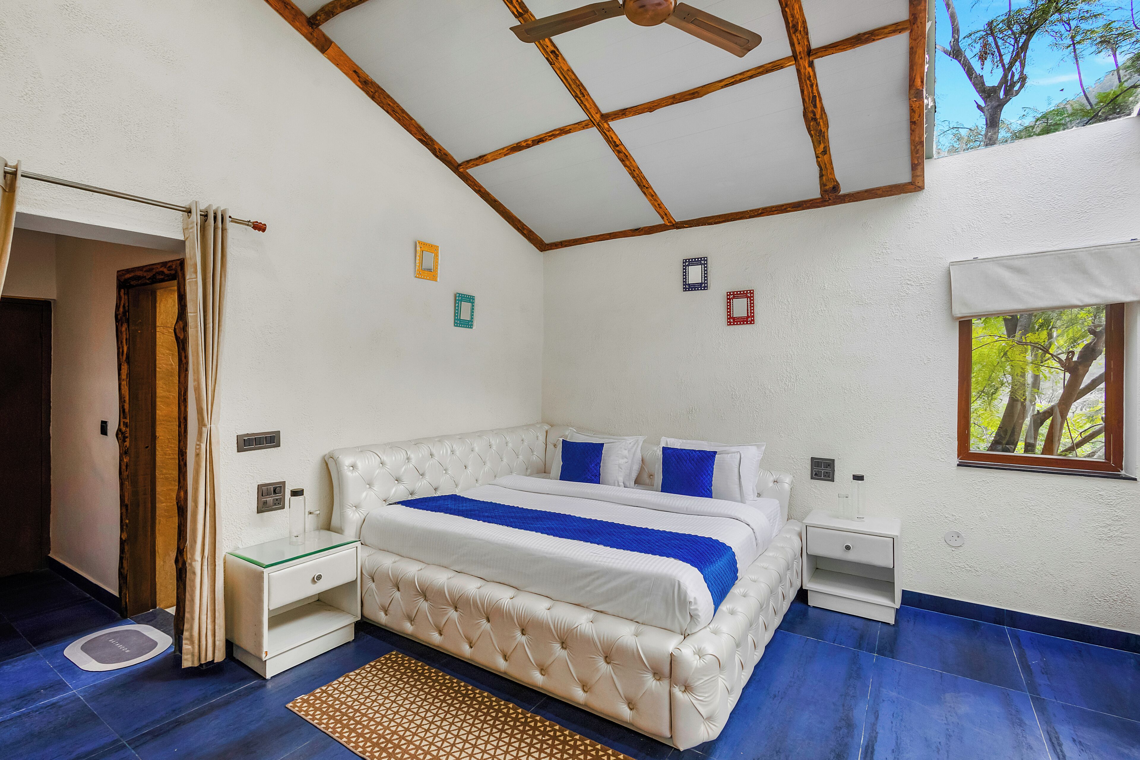 A bedroom with a large bed, wooden ceiling beams, a small bedside table, and a window with a view of trees.
