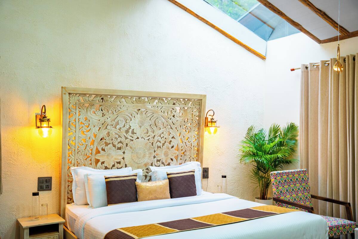 A hotel room with a large bed, ornate headboard, two wall-mounted lights, a potted plant, and a window with a view.