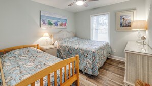 Une chambre avec deux lits, un ventilateur de plafond, une fenêtre avec des stores et une table de chevet en osier.