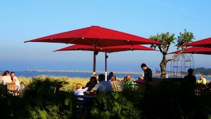 An outdoor dining area with red umbrellas, tables, and chairs, overlooking a body of water. - Vacation home Rügentraum with fireplace and sauna in TOP location (Glowe)