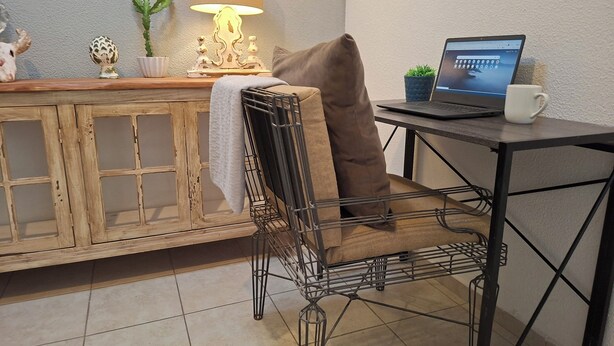 Een stoel met kussen, een laptop op een tafel, een lamp en een kastje met decoratieve objecten.