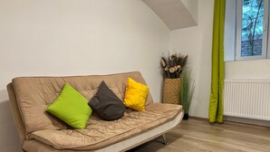 A beige sofa with three cushions in green, black, and yellow, positioned against a white wall with a window and a radiator. - Long Stay 1BR Apt Near Prater Park (Vienna)