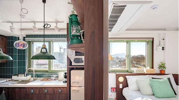 Un espacio habitable pequeño con kitchenette, una cama con almohadas verdes y vistas a la ciudad a través de amplias ventanas.