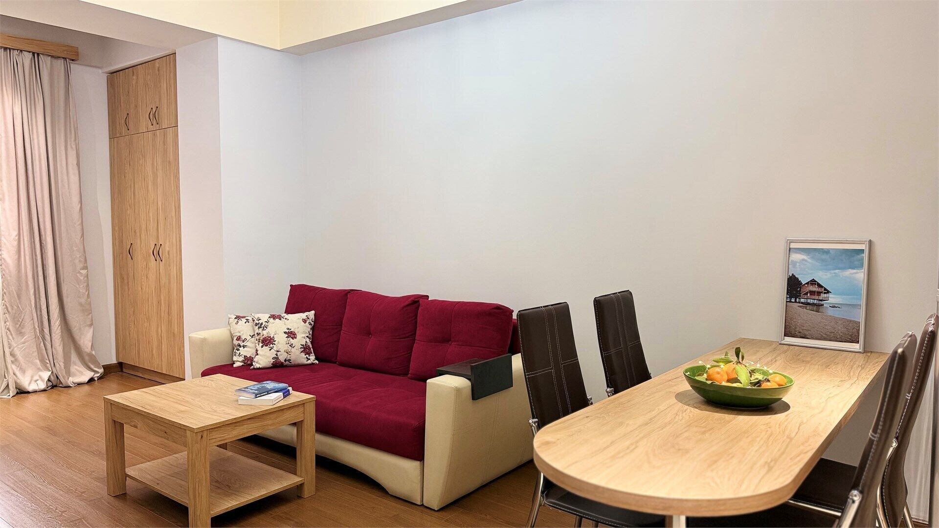 A compact living space with a sofa, a small table, a dining table with chairs, a wooden wardrobe, and a framed picture on the wall.