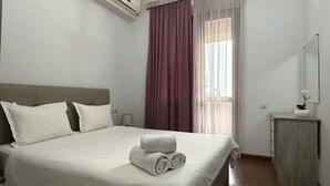 Een netjes opgemaakt bed met witte lakens en gerolde handdoeken, een grijs hoofdbord, een nachtkastje met een lamp en een raam met dunne gordijnen.