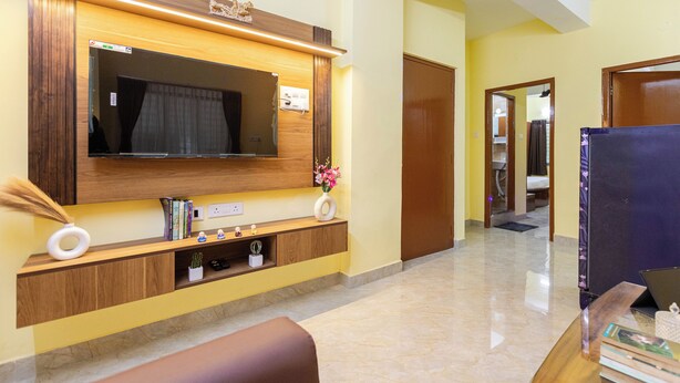 A modern living room with a large flat-screen TV mounted on a wooden wall, a comfortable sofa, and a small coffee table. - City Haven by Jadecaps Nr. Belle Vue Hsp 3bhk (Kolkata)