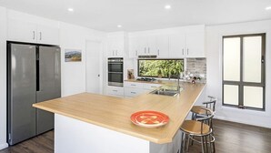 A modern kitchen with white cabinetry, a stainless steel refrigerator, a wooden island with stools, and a large window. - Beautiful House with Mountain and Water Views and Kangaroos. (Long Beach)