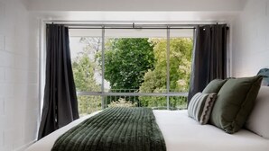 Une chambre avec une grande fenêtre, un lit recouvert d  ’une couverture verte et un balcon donnant sur les arbres.