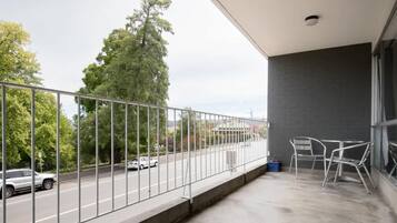 Uma varanda com guarda-corpo de metal, piso de concreto e vista para uma rua com carros e árvores.