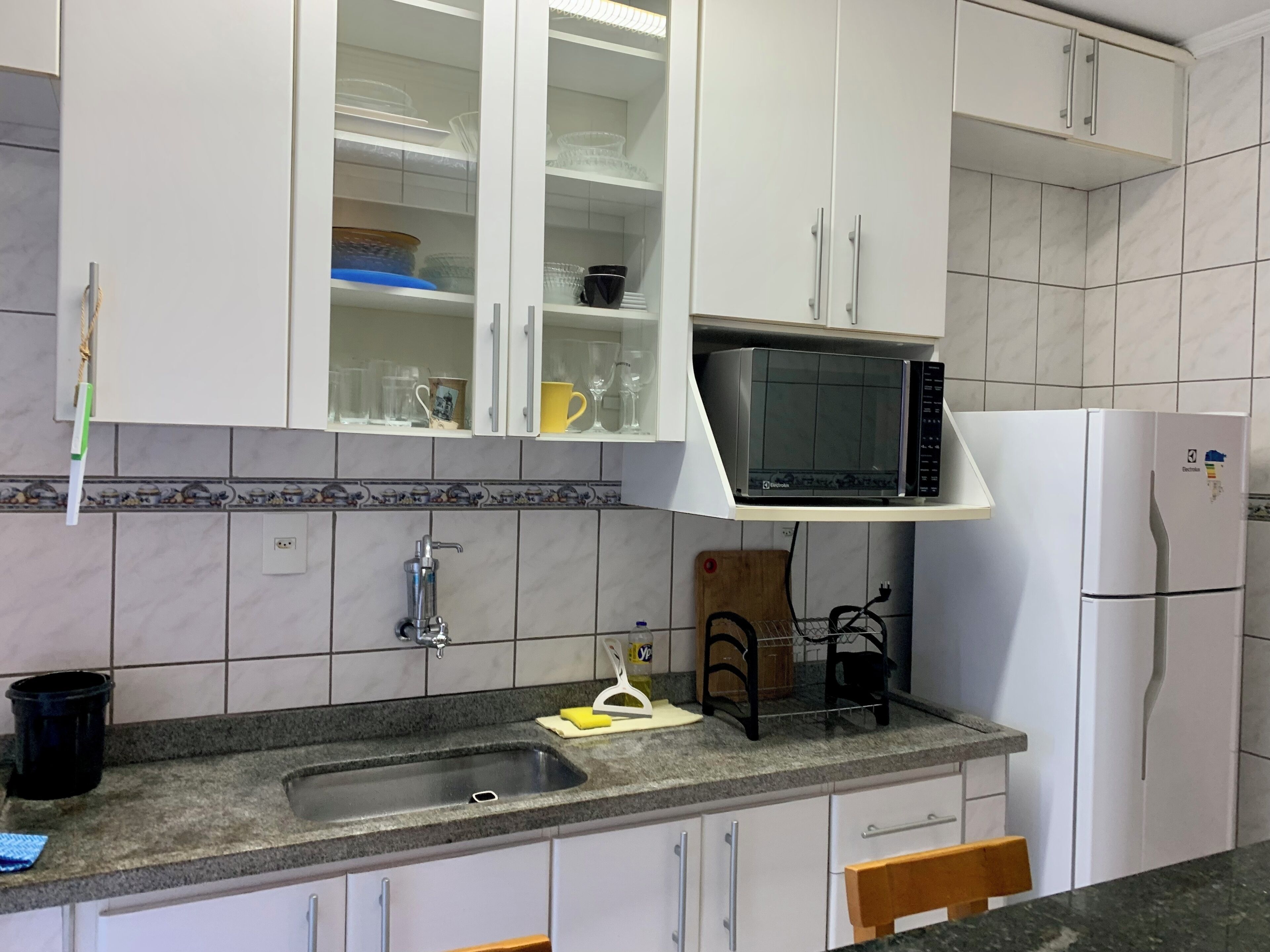 Een keuken met witte kastjes, een inbouwmagnetron, een koelkast, een spoelbak en een houten snijplank.
