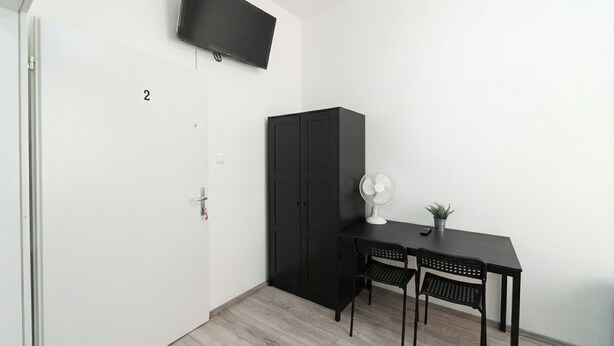 A room with a door labeled '2', a black cabinet, a desk with two chairs, a wall-mounted TV, a standing fan, and a small potted plant. - 15 Min to Vienna Center - Affordable & Simple 5BR Apt with WIFI (Vienna)