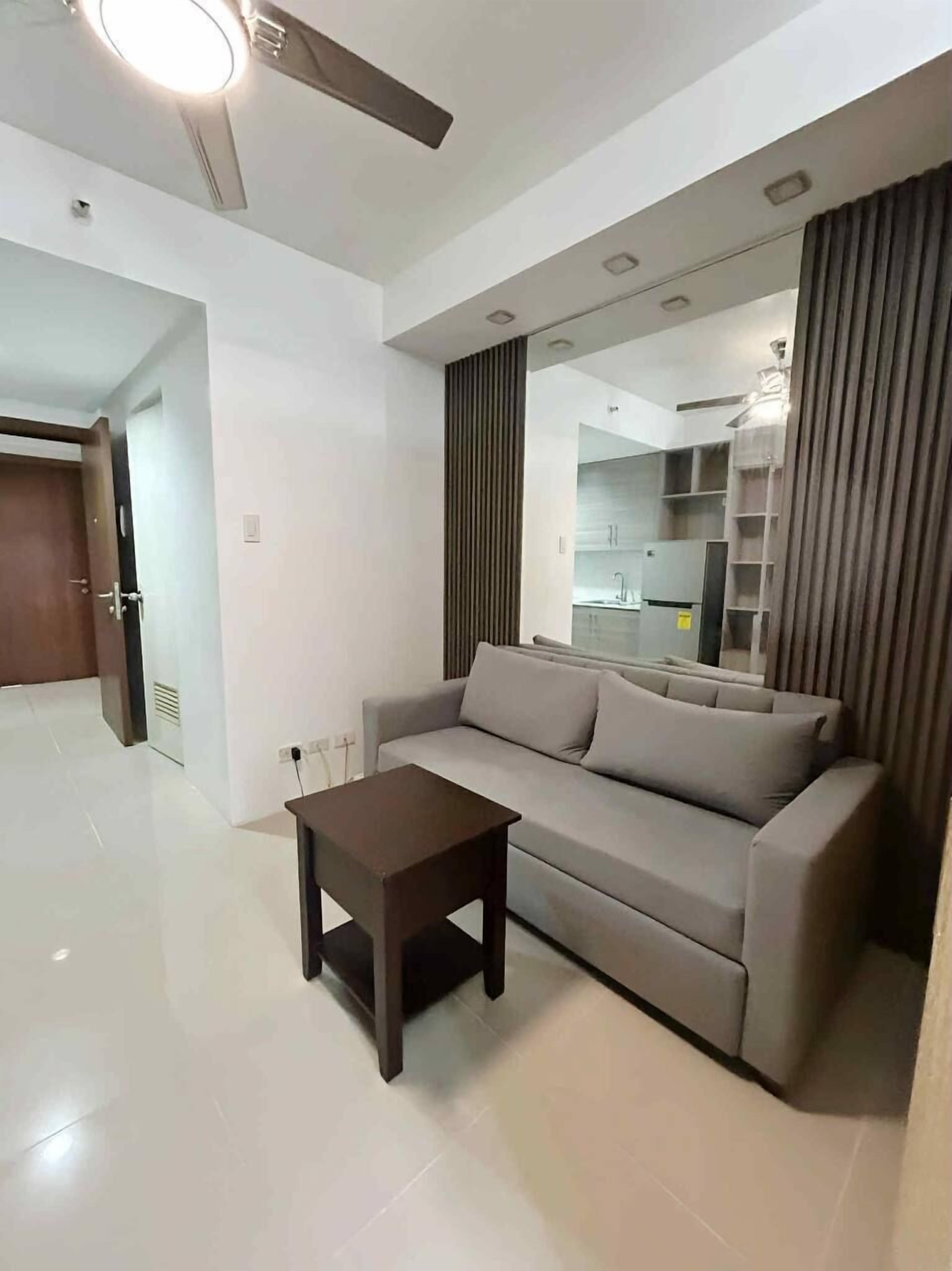A modern living room with a sofa, a small table, a ceiling fan, and a kitchen area visible through a glass partition.