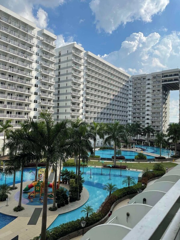 Un hotel con varios pisos, una zona de piscina con palmeras y un área de juegos para niños.
