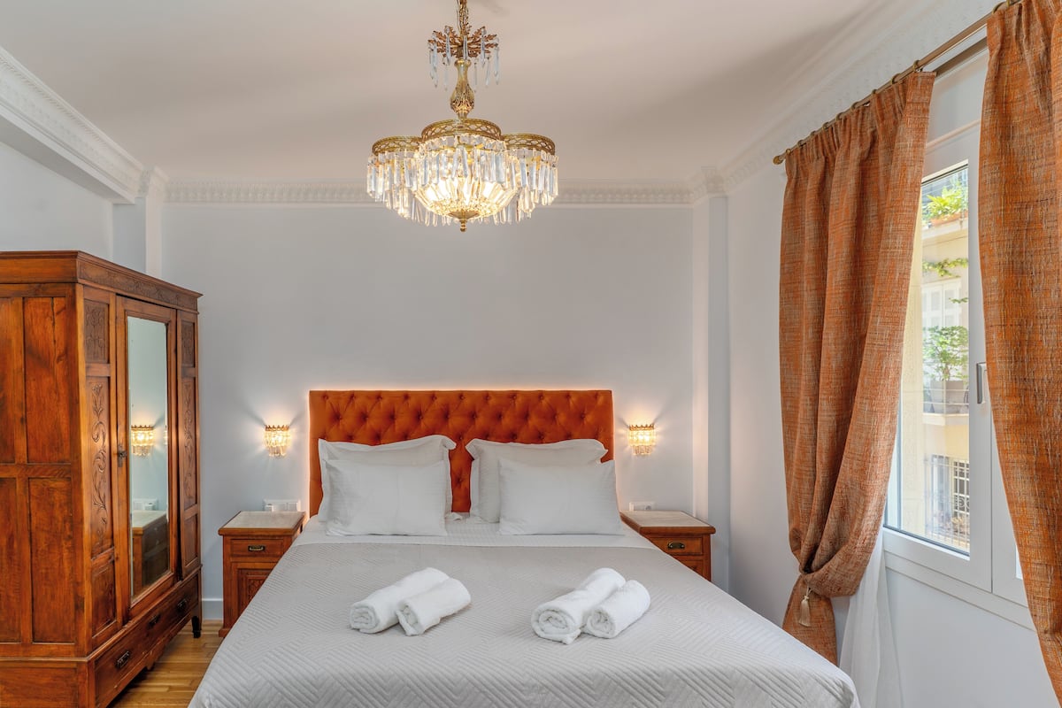 A hotel room with a large bed, a wooden wardrobe, a chandelier, and a window with curtains.