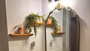 A bathroom with a white marble sink, gold faucets, and a large mirror with an ornate frame. There are wooden shelves above the mirror holding potted plants and decorative items. - Welcome to my My Cozy Mushroom cottage ! (Big Bear)