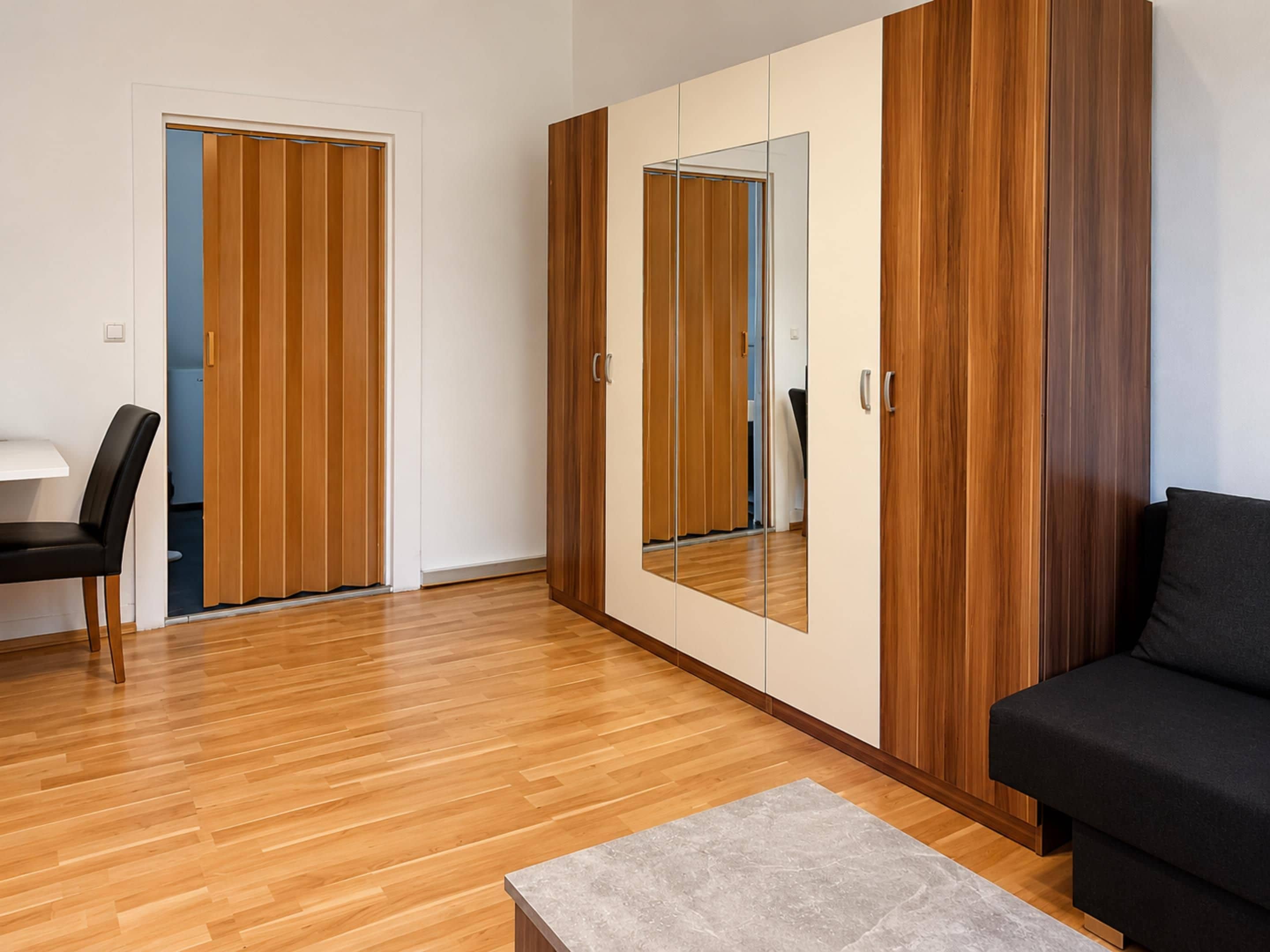 Ein modernes Zimmer mit Holzböden, einem schwarzen Sofa, einem kleinen Tisch und einem Schrank mit Spiegel.