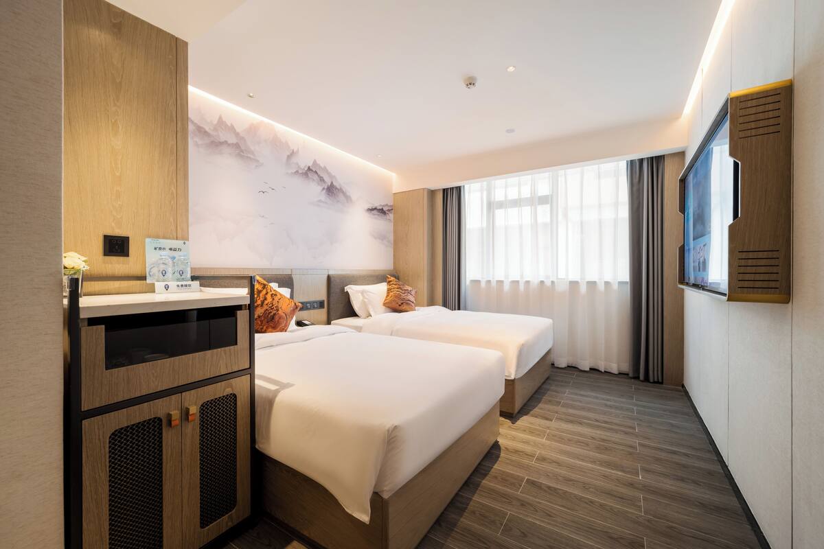 Een hotelkamer met twee bedden, een houten garderobe, een tv aan de muur en een groot raam met gordijnen.