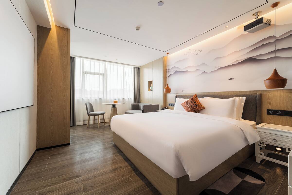 Een moderne hotelkamer met een groot bed, een bureau en een stoel. Er is een raam met gordijnen, een wandprojector en een plafondlamp.