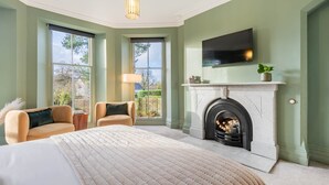 A bedroom with a large bed, a fireplace, a TV mounted above it, and two chairs. - Loughrigg at Lipwood House (Windermere)