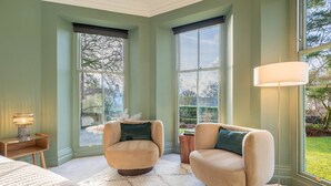 A bedroom with two armchairs, a bed, a nightstand, a lamp, and large windows with a view of greenery. - Loughrigg at Lipwood House (Windermere)