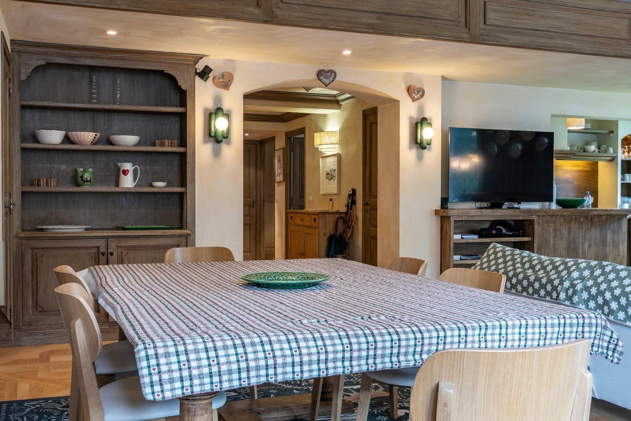 Un comedor con mantel a cuadros, sillas de madera, un plato verde y un televisor al fondo.