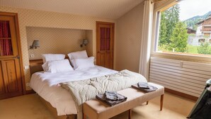 A bedroom with a bed, a bench, two wall-mounted lamps, and a window with a view of buildings and trees. - Spacious 4BDR/9P apt at the foot of the ski slopes (Megève)