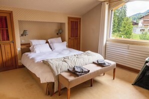 Une chambre à coucher comprenant un lit, un banc, deux lampes fixées au mur et une fenêtre donnant sur des immeubles et des arbres.