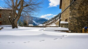Une rue enneigée, avec un bâtiment en pierre, un arbre et des montagnes en arrière-plan.