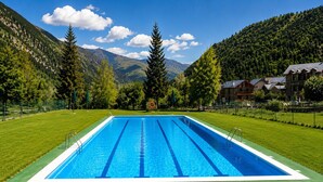 Une piscine rectangulaire à l’eau d’un bleu limpide, entourée de pelouse verdoyante et d’arbres, avec des montagnes en toile de fond.