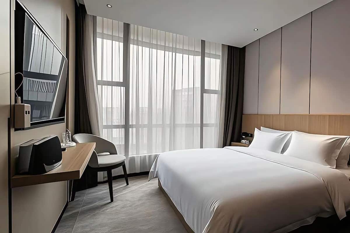 Una habitación de hotel moderna con una cama grande, un escritorio con una silla, un televisor y un ventanal con cortinas.