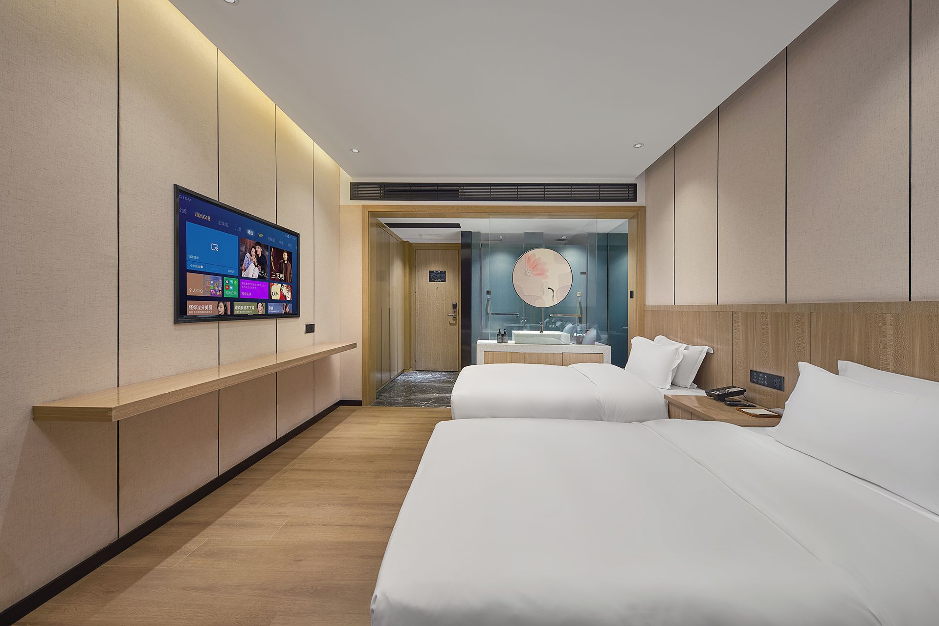 A hotel room with two beds, a TV mounted on the wall, a bathroom with a mirror and sink, and a hallway leading to another room.