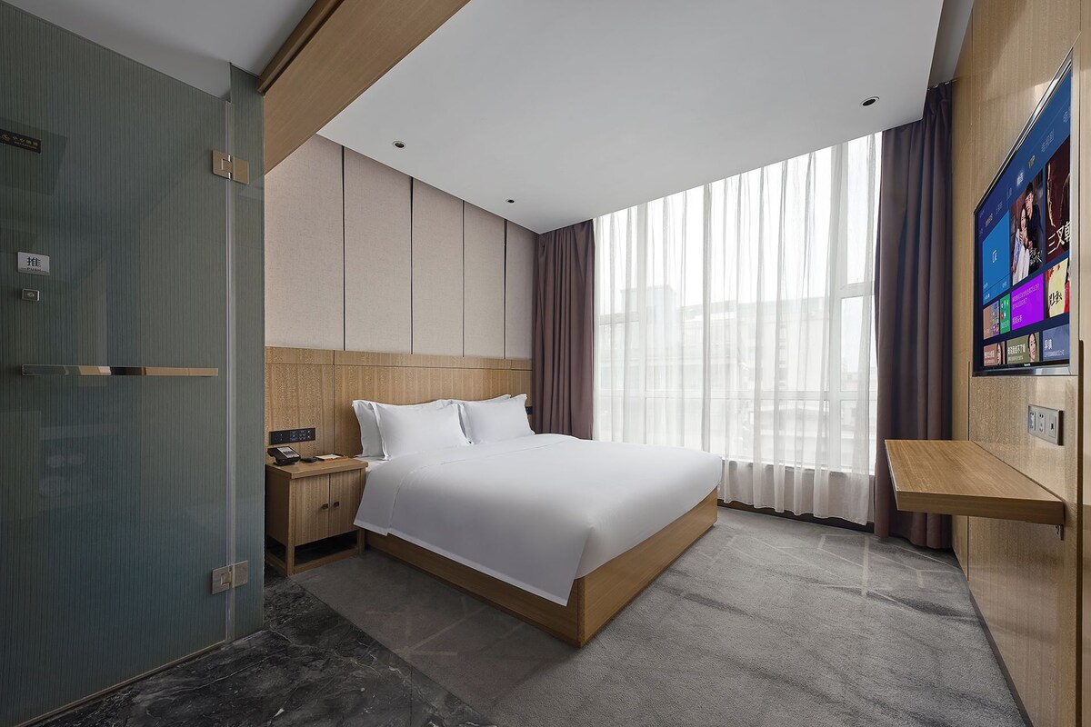 Una habitación de hotel moderna con una cama grande, una mesita de noche, un escritorio y un ventanal con cortinas.