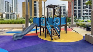 A playground with a slide and climbing structure, surrounded by a colorful play area and modern buildings in the background. - Superhost Modern 1BR in Azizi Riviera (Dubai)