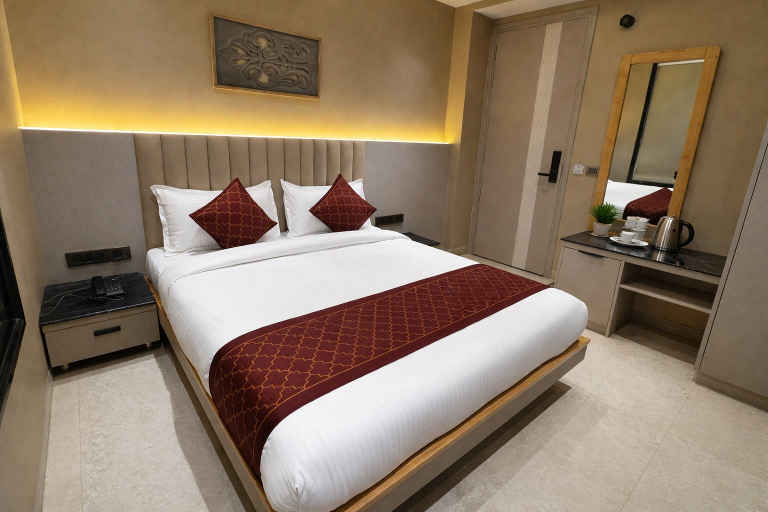 A modern hotel room with a large bed, a bedside table with a phone, a small desk with a mirror, and a coffee station.
