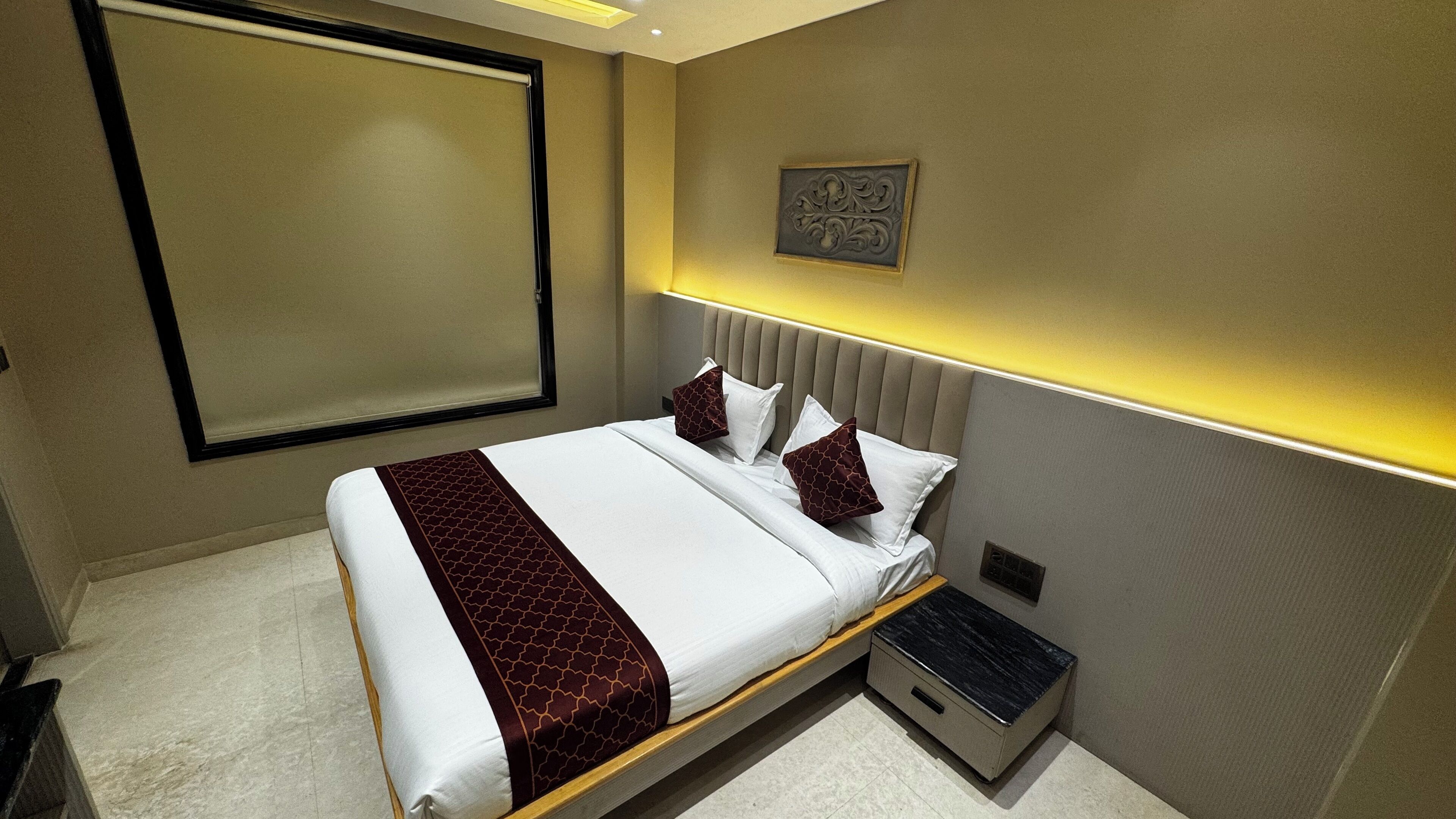 Habitación de hotel con cama, mesita de noche, un armario pequeño, una obra de arte en la pared y una ventana con persiana.