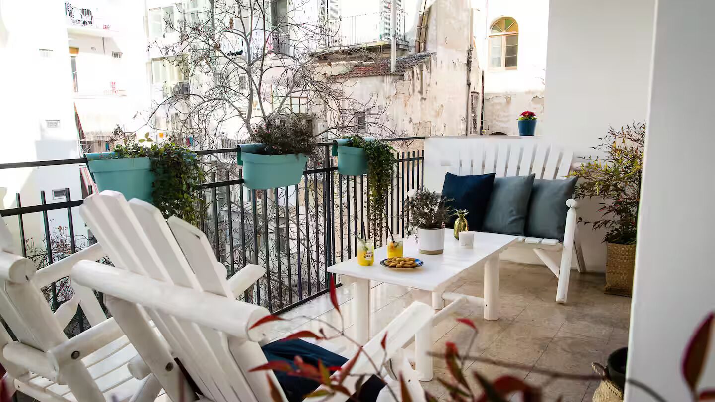 A balcony with white chairs and a table, potted plants, and a view of buildings and a river.
