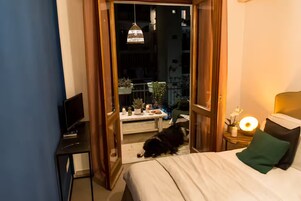A hotel room with a bed, a desk with a laptop, a bedside table with a lamp, and a view of the outdoor balcony area.