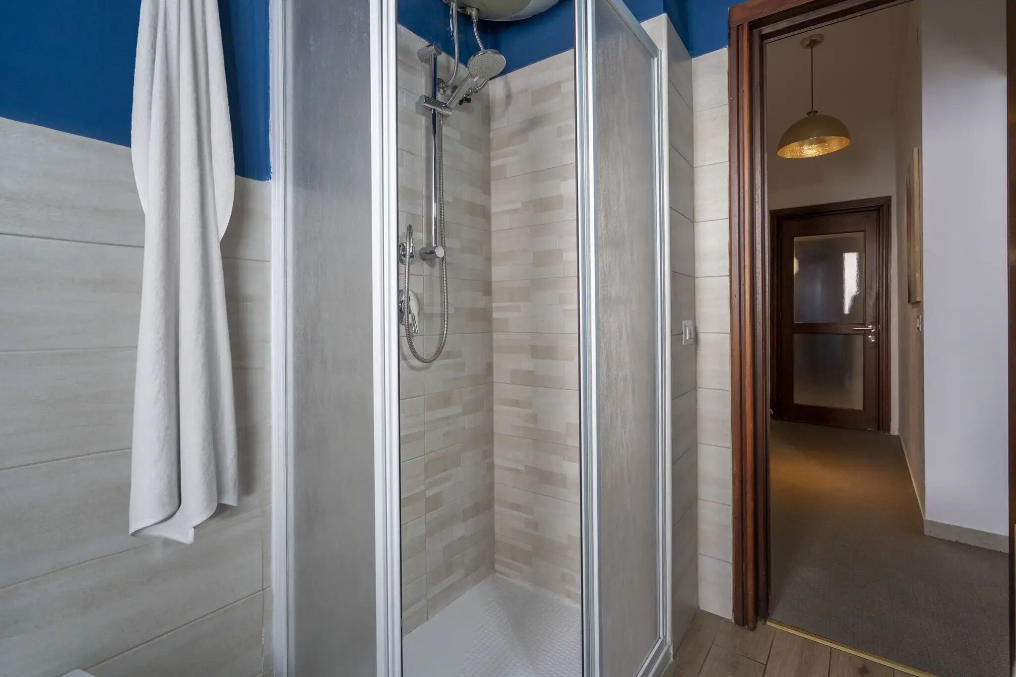 A modern bathroom with a glass shower enclosure, a white towel hanging, and a wooden door leading to a hallway.