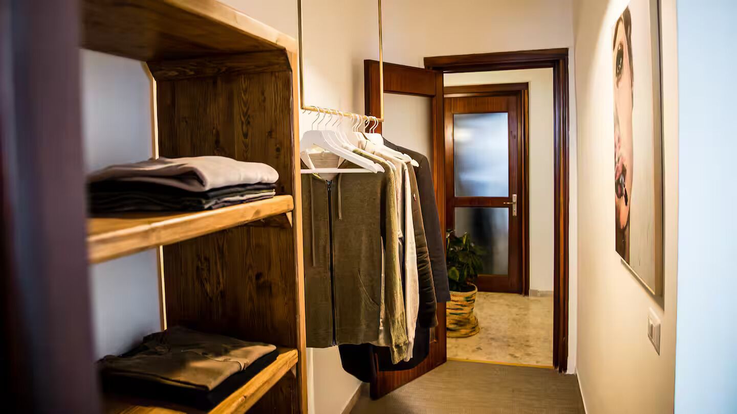 A narrow hallway with wooden shelves on the left, a clothing rack in the center, and a door at the end.