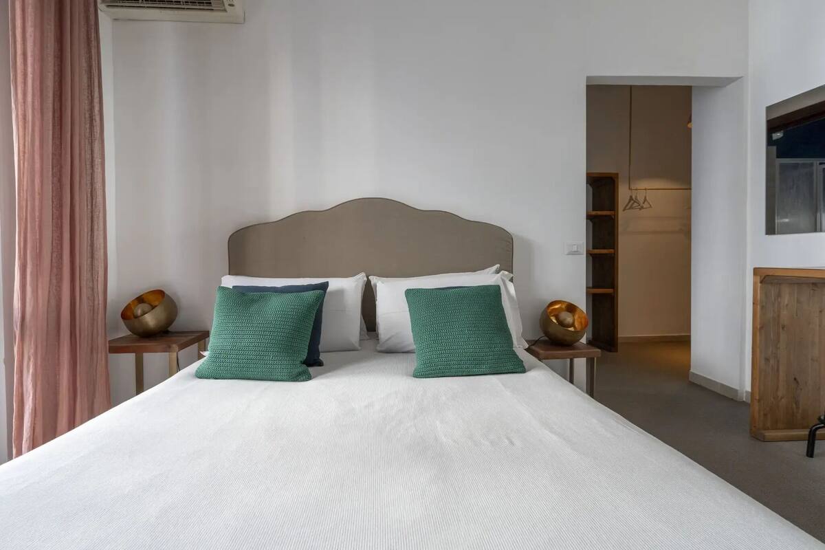 A neatly made bed with a white duvet and two green pillows, a wooden headboard, and a small bedside table with a bowl.