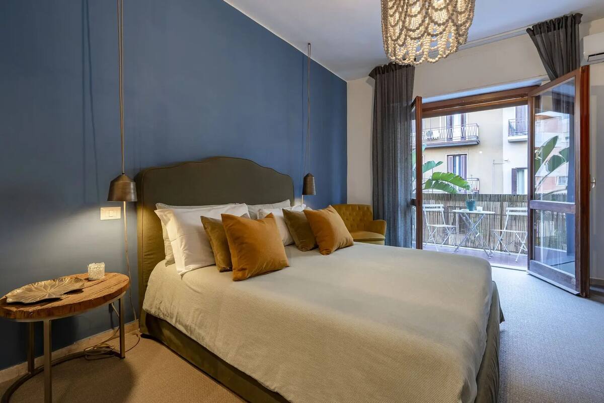 A hotel room with a large bed, a small round table, a hanging light fixture, and a balcony with outdoor furniture.