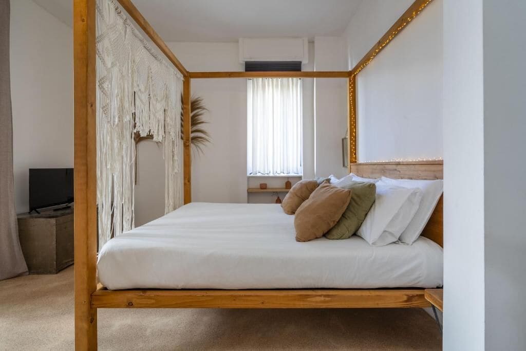 Een slaapkamer met een houten hemelbed, een televisie, een raam met gordijnen en een plant.