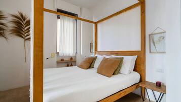 Un lit à baldaquin avec du linge de lit blanc et de nombreux oreillers, une tête de lit en bois, une table de chevet avec une bougie et une œuvre d’art botanique encadrée accrochée au mur.