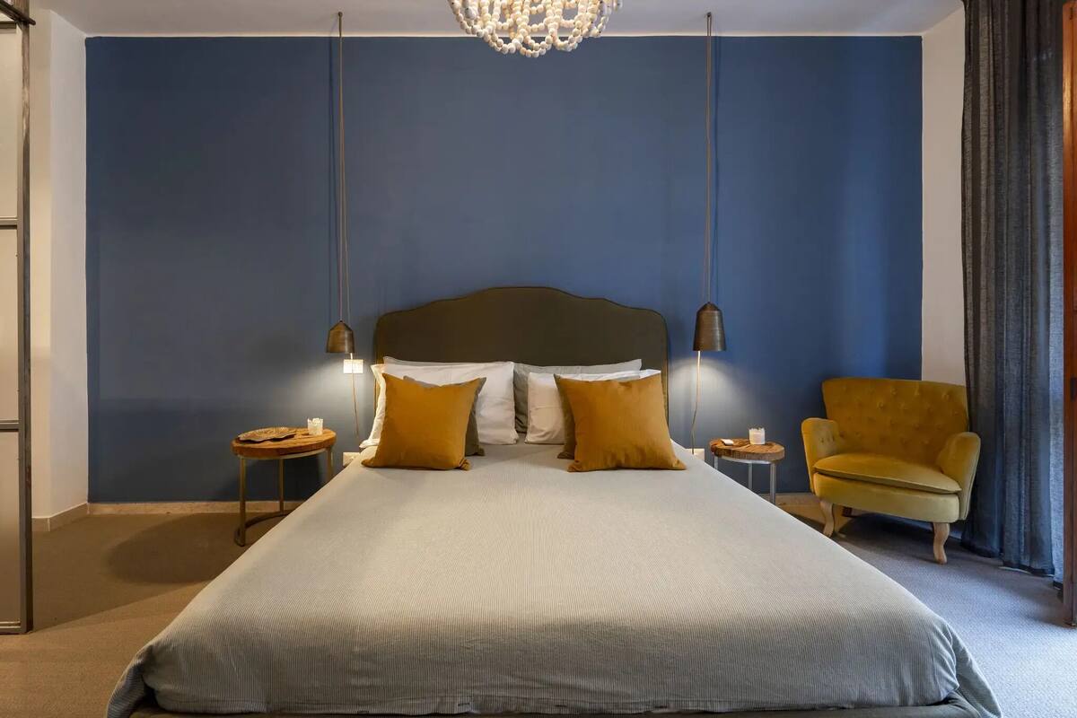 A neatly made bed with a headboard, two bedside tables with lamps, a yellow armchair, and a chandelier.