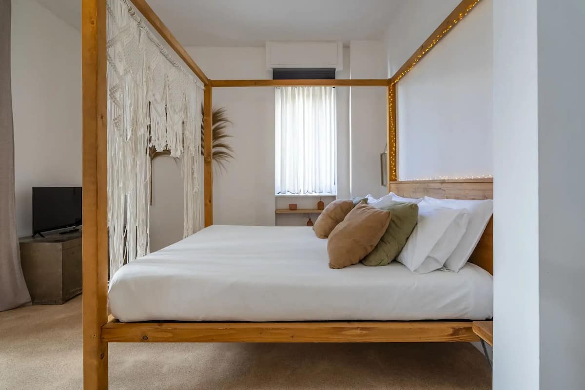 A bedroom with a wooden four-poster bed, a television, a window with curtains, and a small shelf with decorative items.